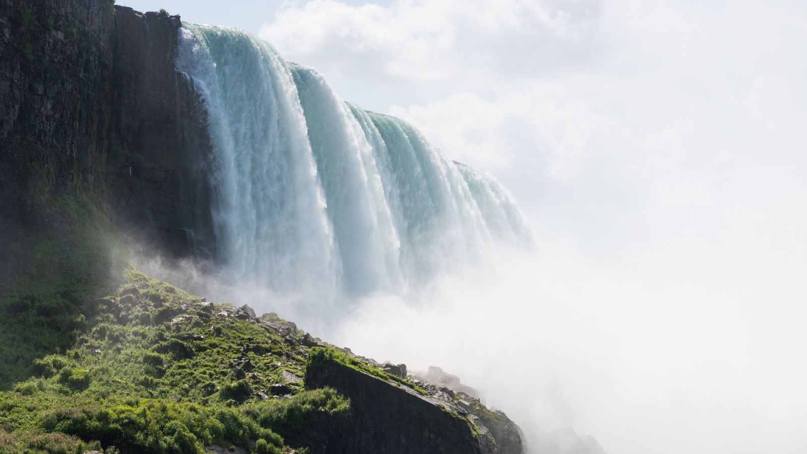 Waterfalls pour over a rocky cliff, surrounded by mist and lush greenery beneath a cloudy sky.