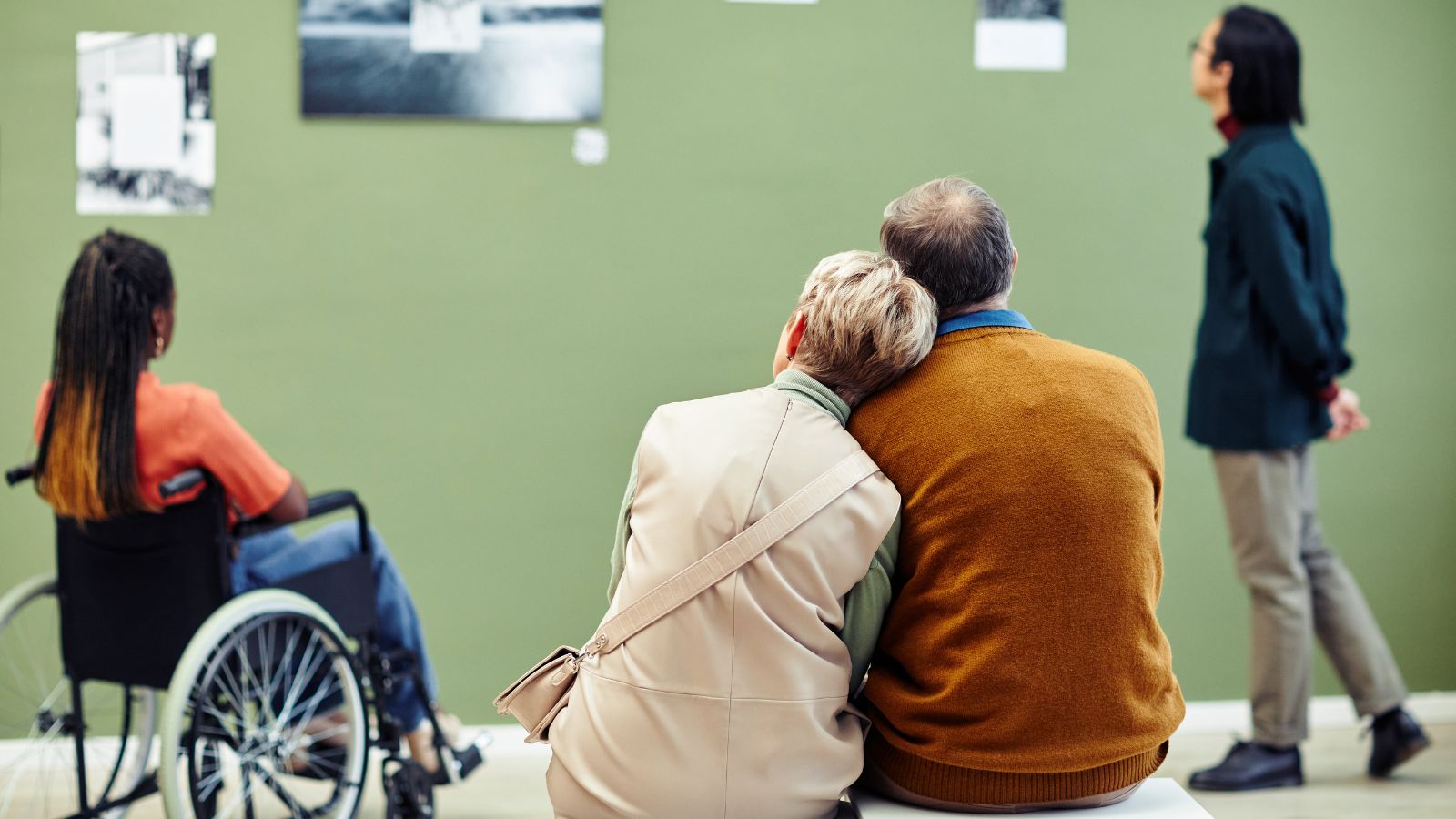Three people look at photos on a green wall; nearby, a woman in a wheelchair and an older couple sit close together.