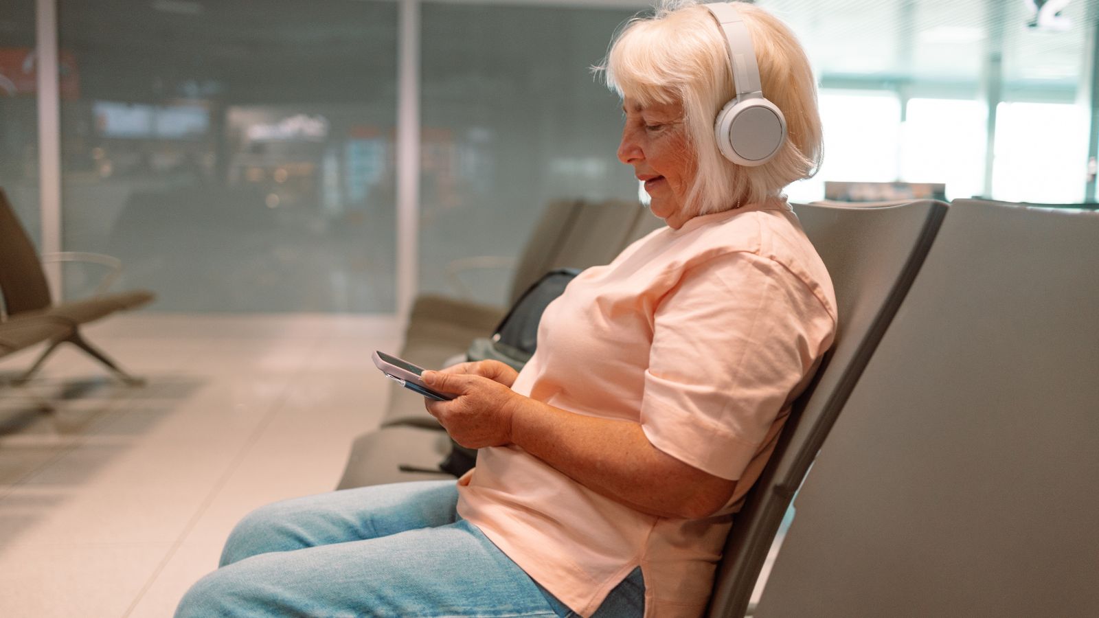 Older woman in an airport lounge with headphones, looking at her phone, empty seats next to her.