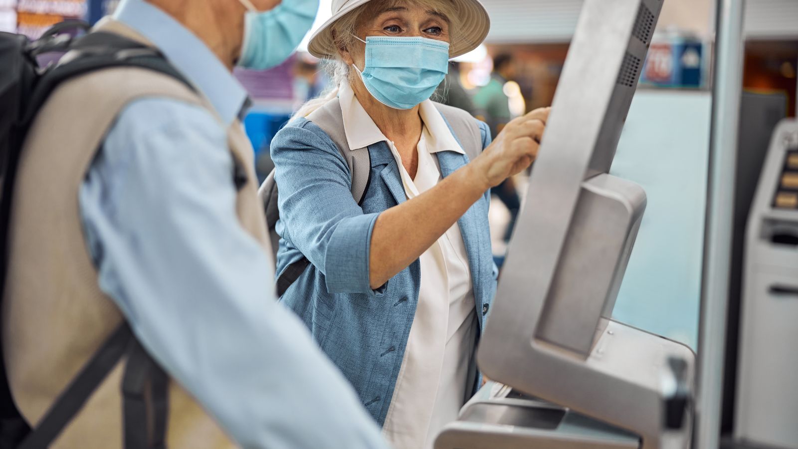 Two masked individuals use a self-service kiosk, with one person touching the screen.