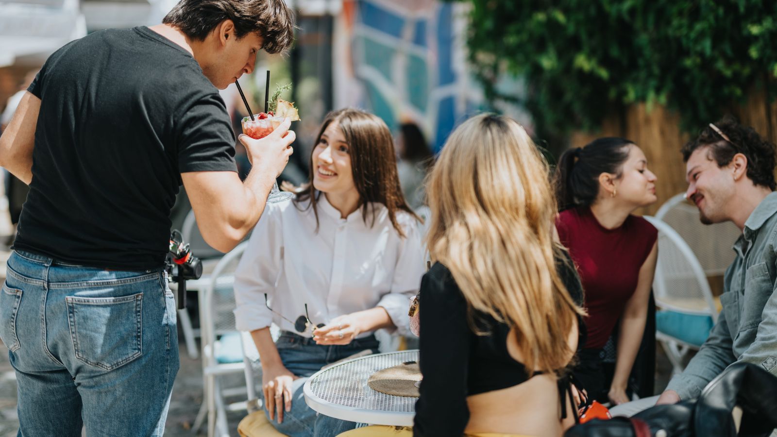 Four people sit at an outdoor café table chatting and smiling as a man stands nearby holding a garnished drink with a straw.