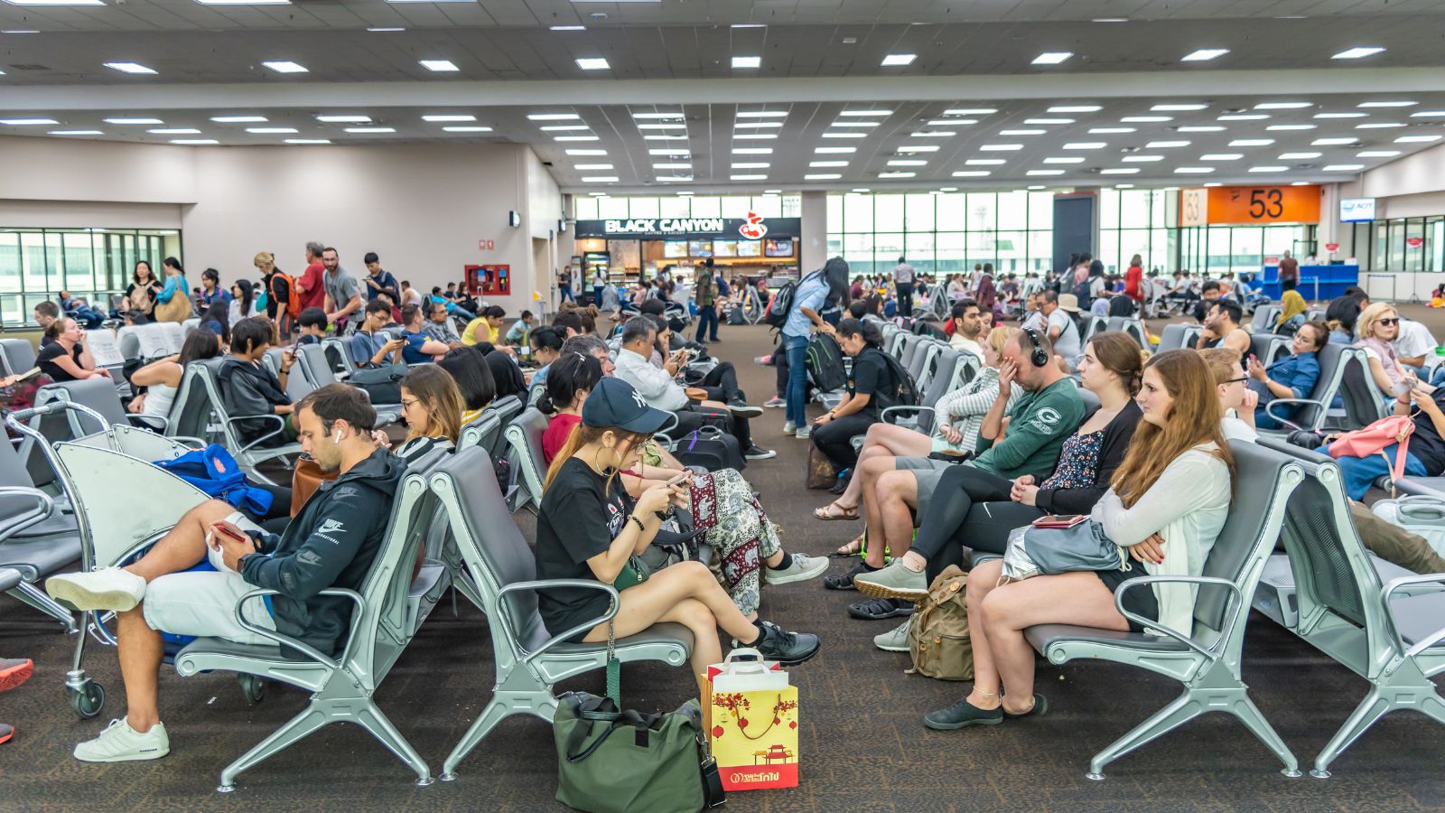 Passengers wait among rows of chairs and luggage at a crowded airport gate, with overhead signs and a café in the background.