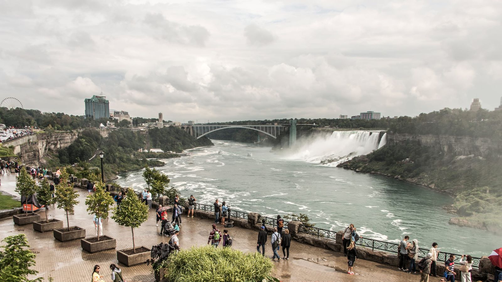 Niagara Falls with mist, flowing river, bridge in the background, and tourists on a riverside promenade in the foreground.
