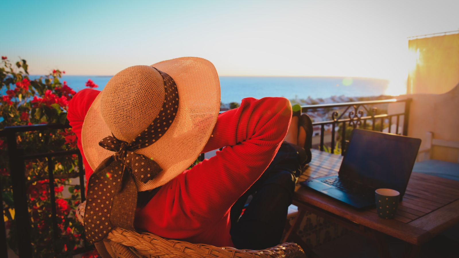 Person in a wide-brimmed hat relaxes on a sunset balcony by the sea, with a laptop and coffee cup on the table nearby.