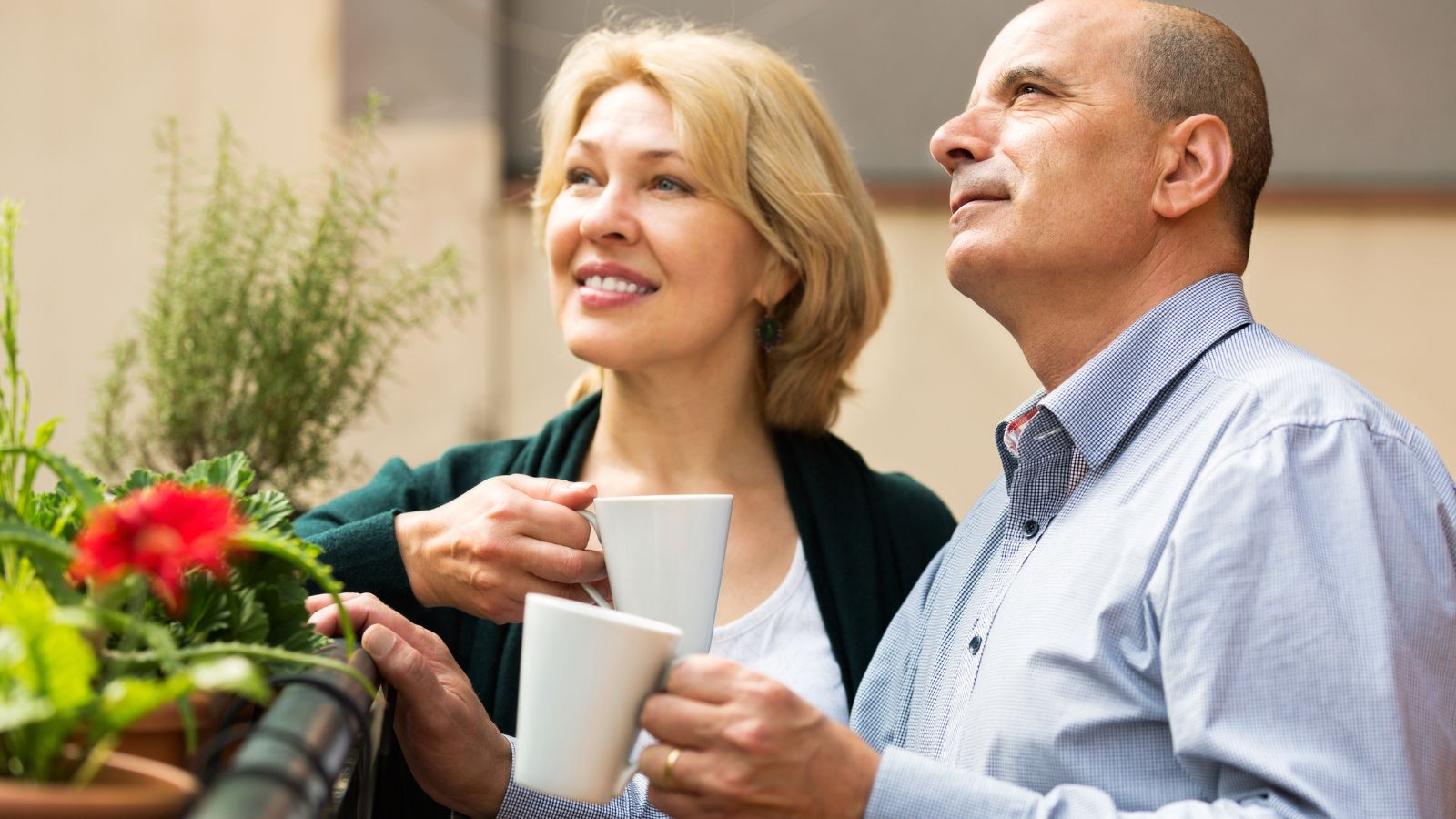 Two adults with white mugs stand by outdoor potted plants, smiling as they look upward.