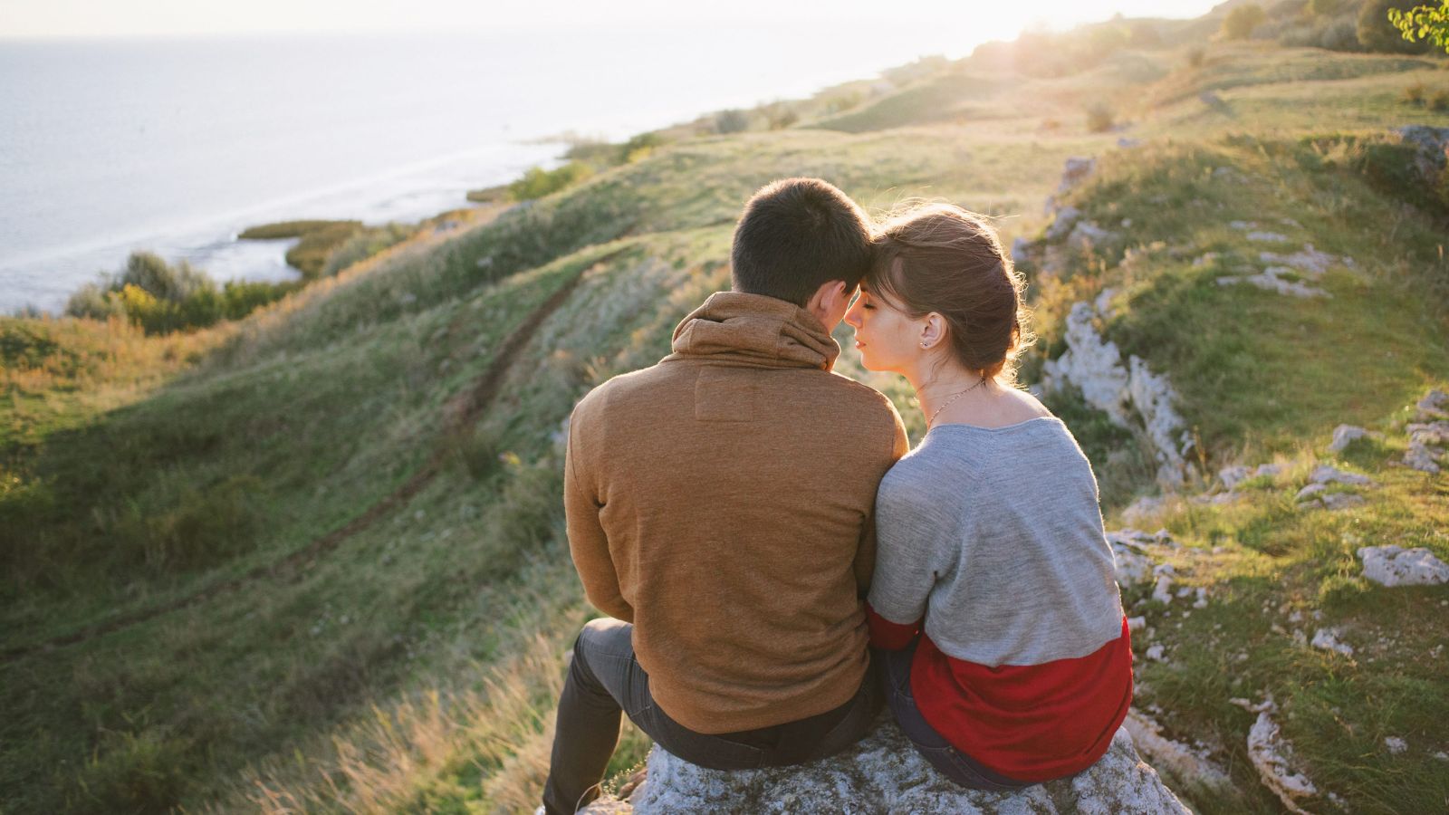 Two people sit closely on a rock, gazing over grassy land and water at sunset.