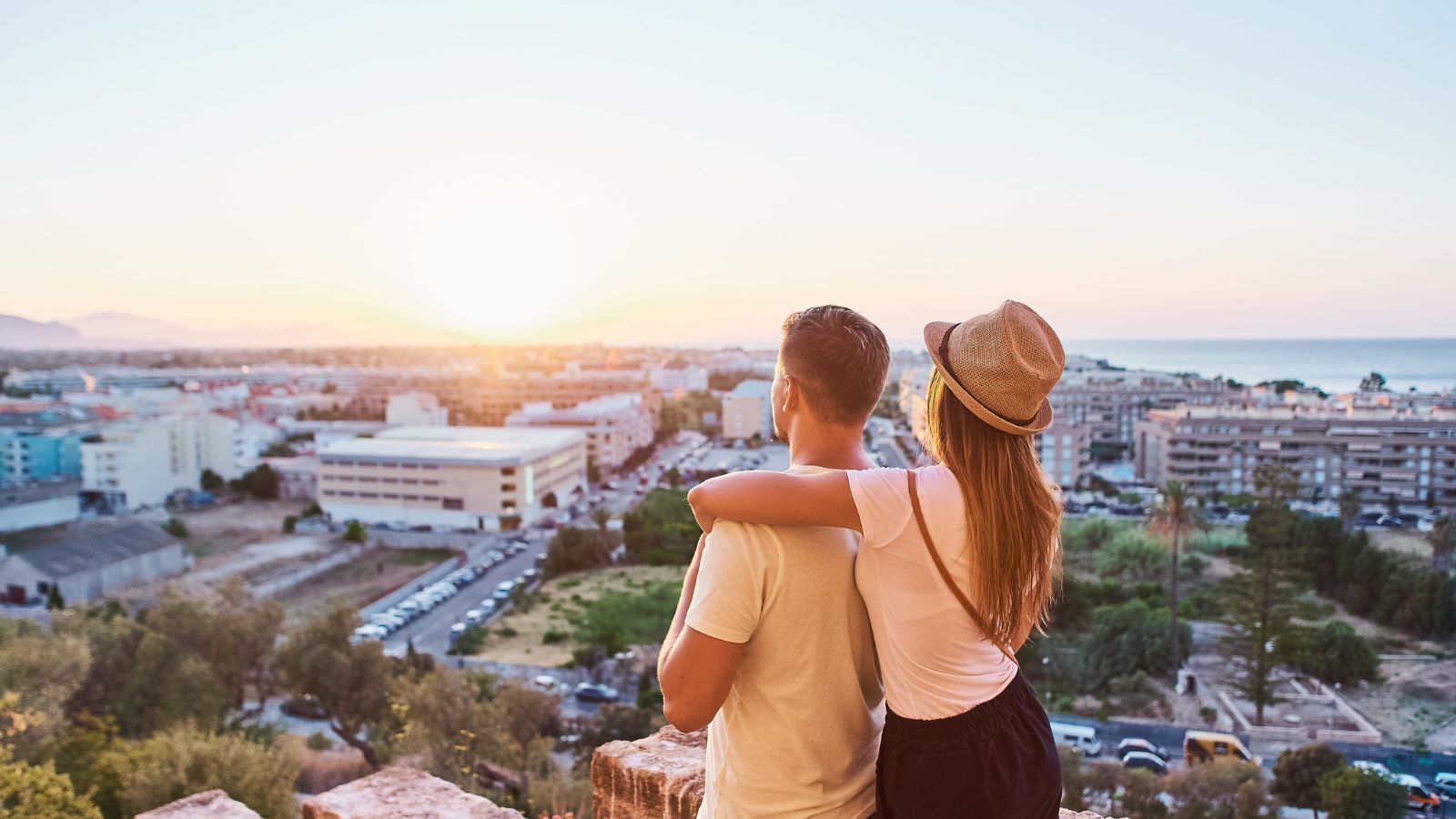 A couple stands on a hill at sunset, the woman’s arm around the man as they face the cityscape and setting sun together.