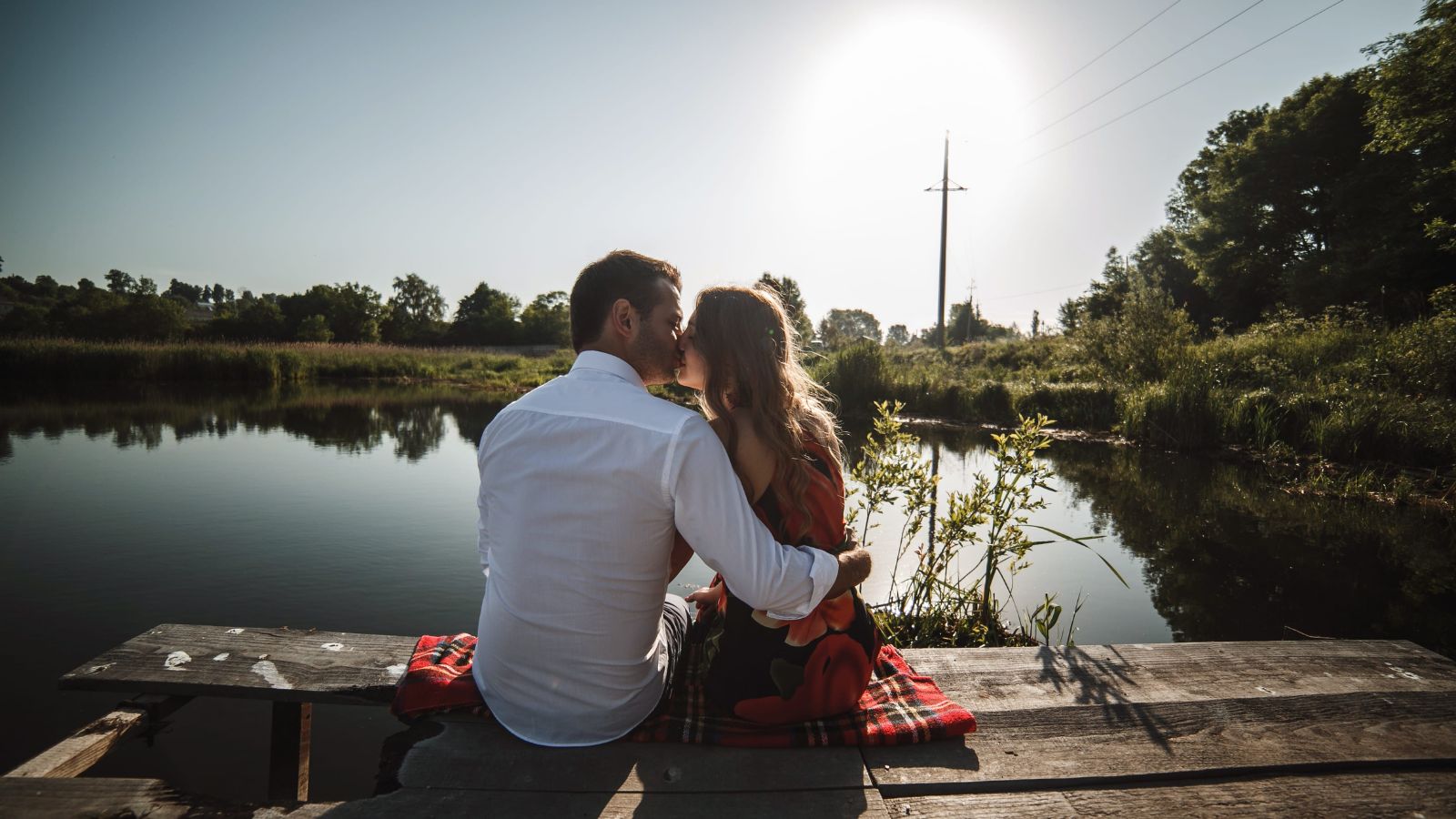 A couple sits closely on a wooden dock by a calm lake at sunset, surrounded by trees and greenery.