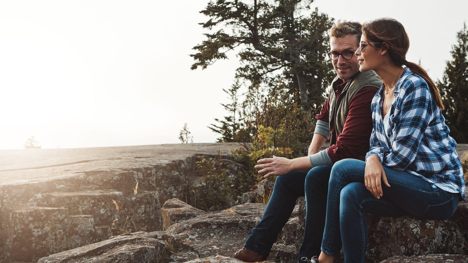 Two people sit on rocky ground outdoors, talking face-to-face with trees and sunlight in the background.