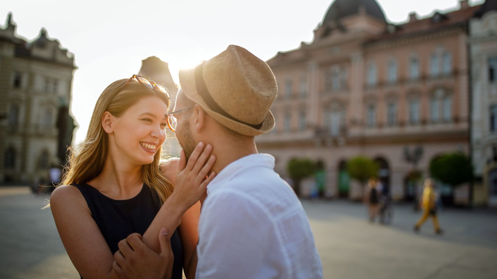 A woman smiles, touching a man's face as they stand close in a sunlit city square with historic buildings behind them.
