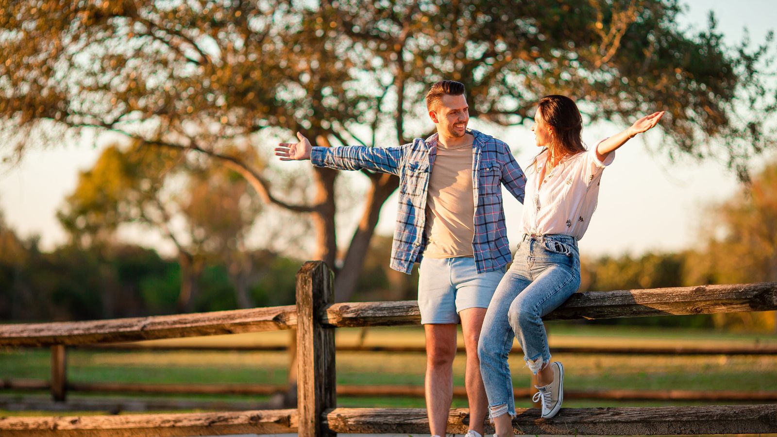 A man and woman with outstretched arms sit and stand on a wooden fence in a sunny park, looking at each other.