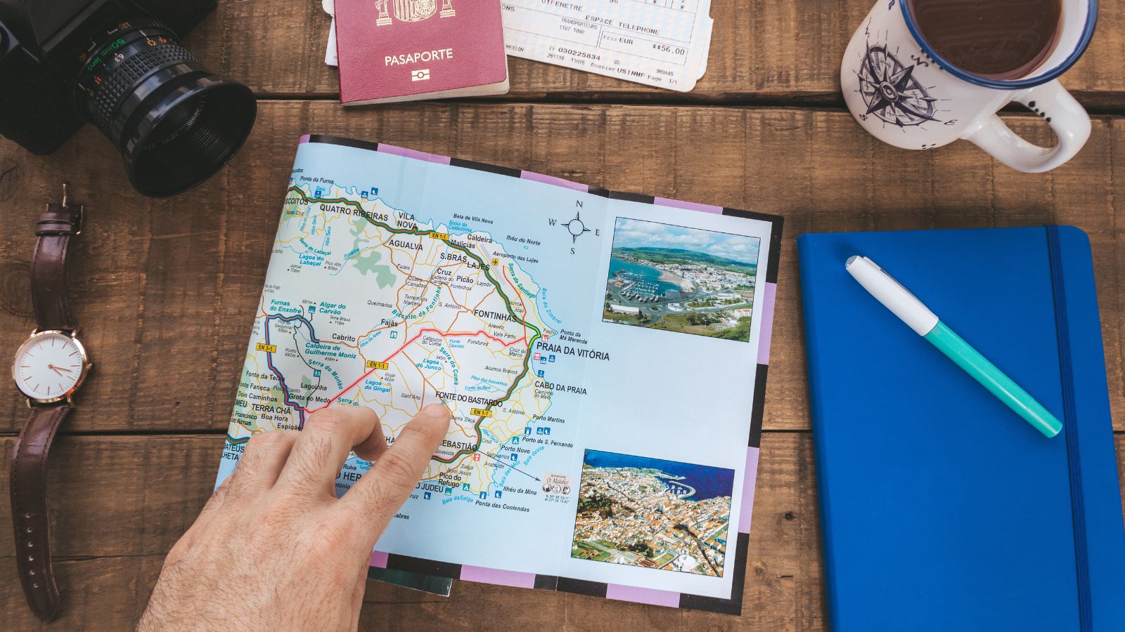 A hand points to a map on a desk surrounded by a watch, camera, passport, boarding pass, coffee mug, notebook, and pen.