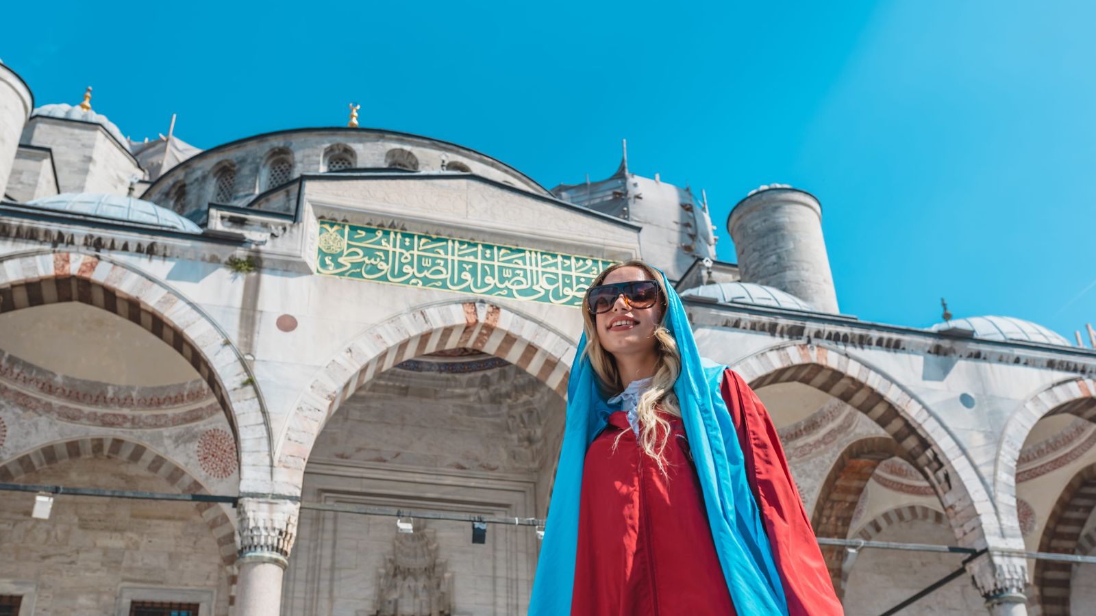 A woman in sunglasses and a headscarf stands before a historic mosque with arched entrances and Arabic script above them.