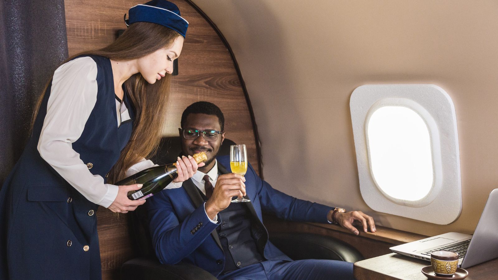 A flight attendant pours champagne for a suited man seated in first class next to a laptop and coffee cup.