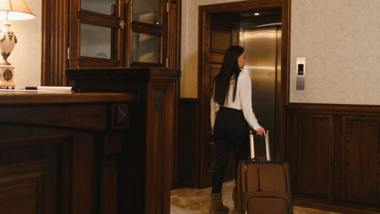 A woman in business attire with long hair pulls a suitcase toward an open elevator in a wood-paneled hotel lobby.