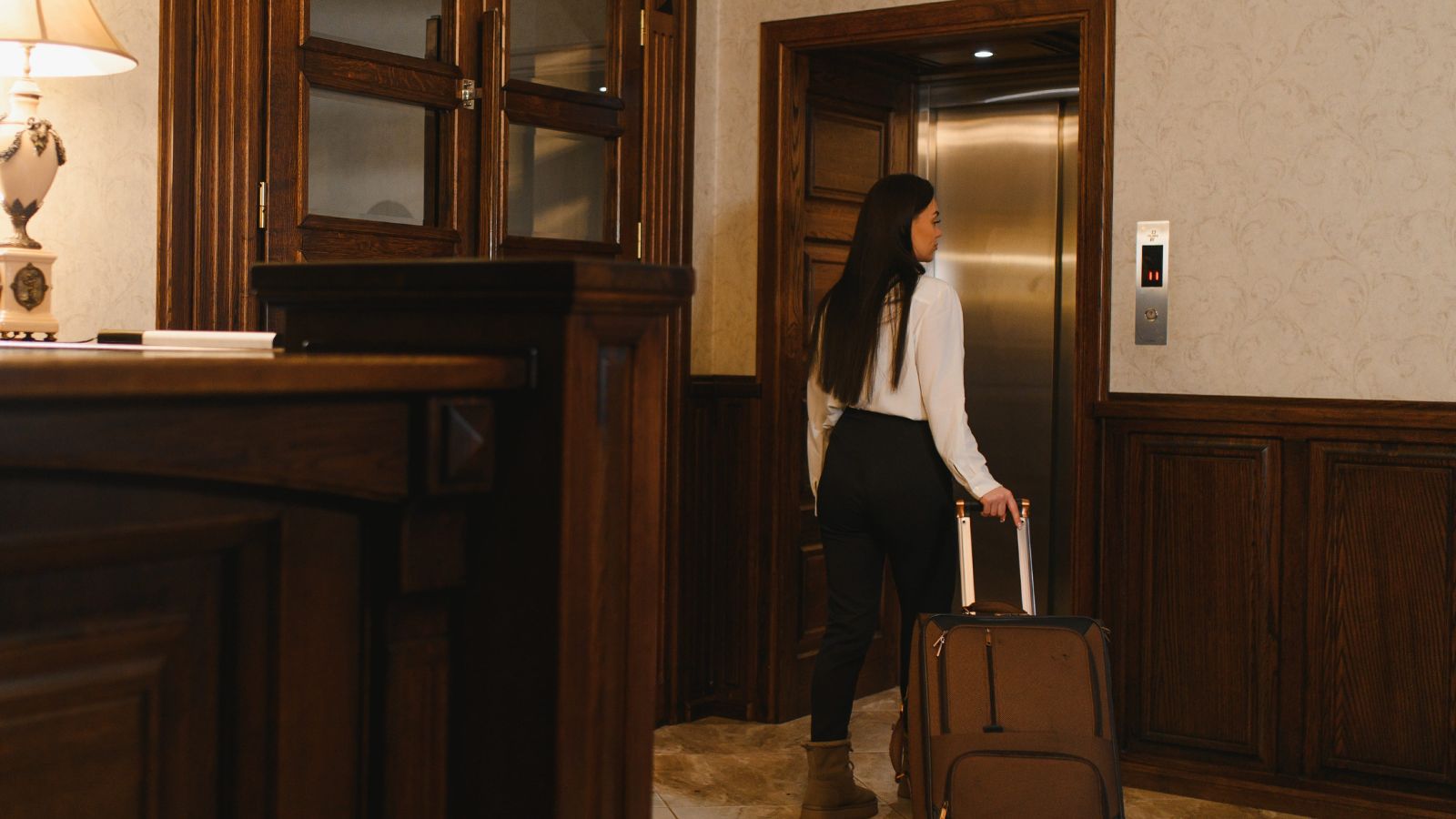 A woman in business attire with long hair pulls a suitcase toward an open elevator in a wood-paneled hotel lobby.