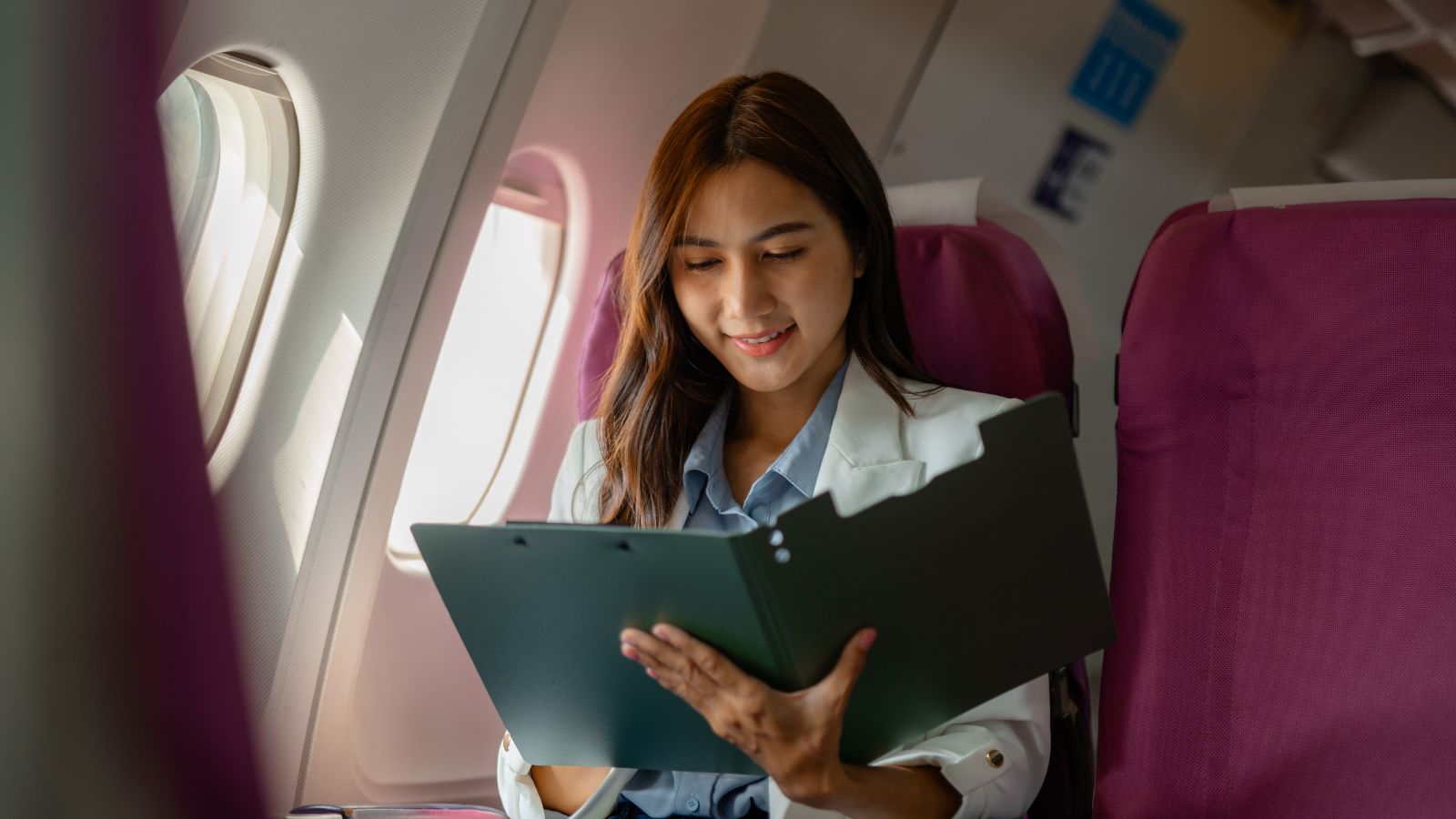 A woman reads documents from a folder while seated by the window on an airplane.
