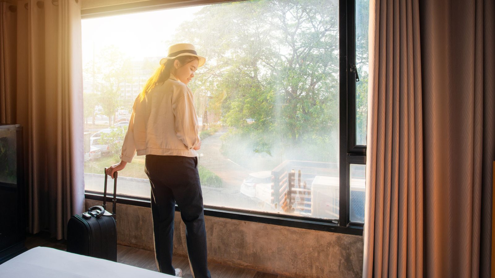 Person with a suitcase stands by a hotel room window, gazing out at trees and sunlight.