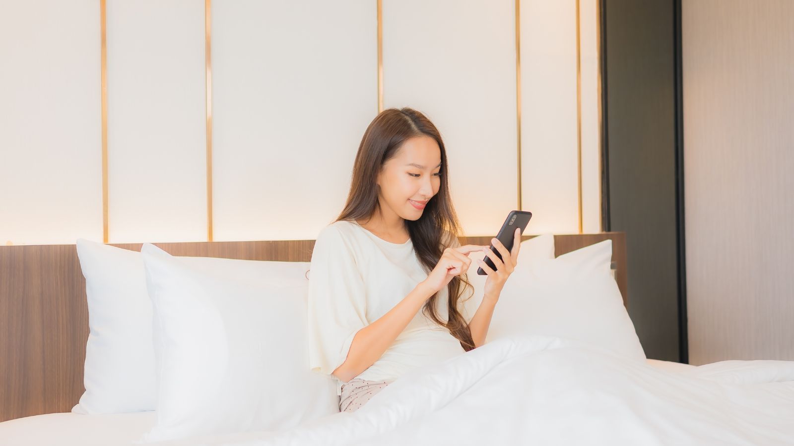 A photo of a woman checking something in her phone in the hotel bed.