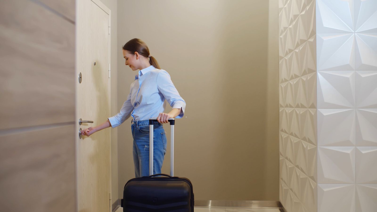 Woman in a blue shirt and jeans stands by a door with a suitcase in a modern hallway with textured white walls.