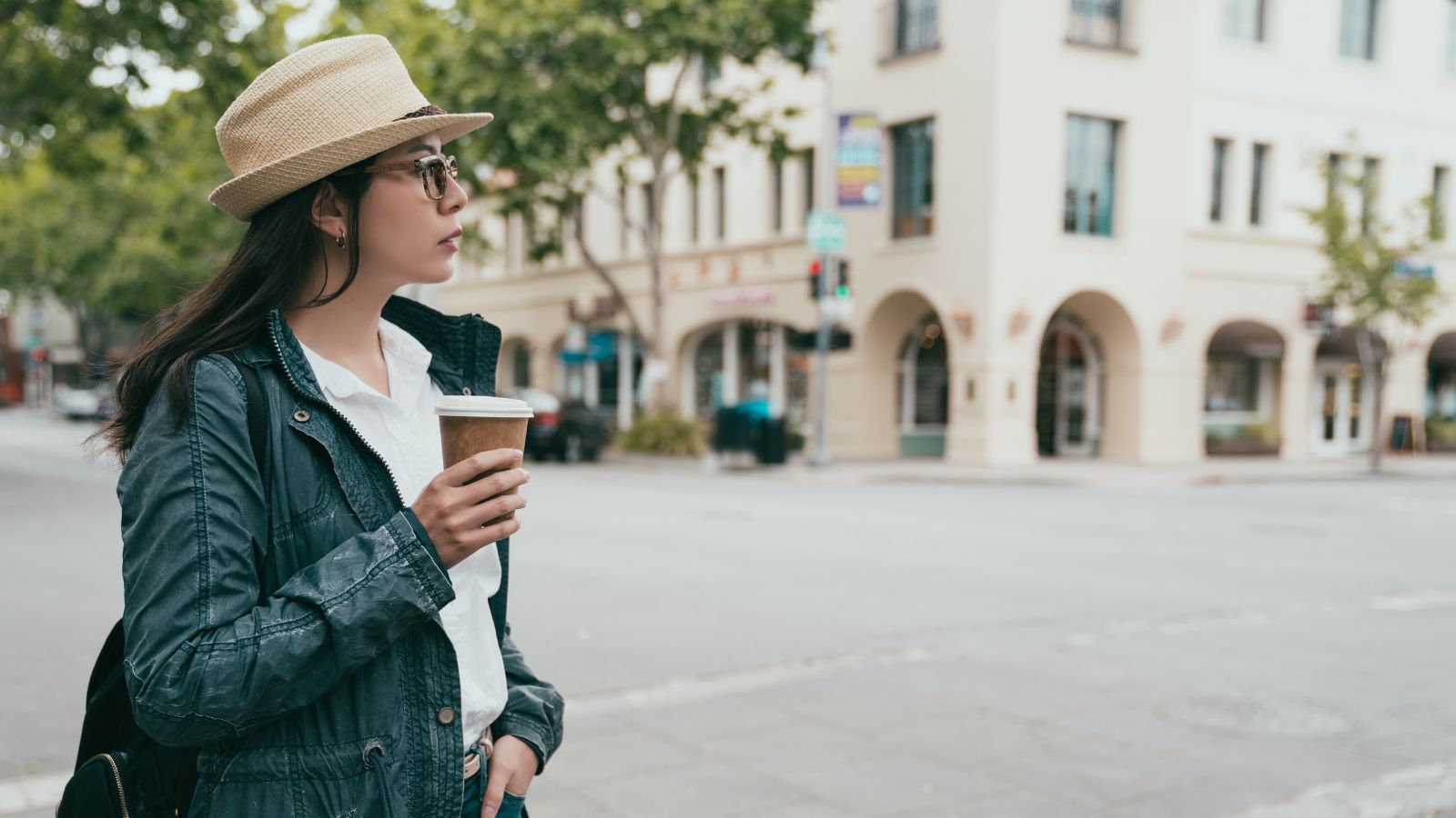 A woman in a hat and glasses stands at a city street corner, holding coffee and looking to the side.