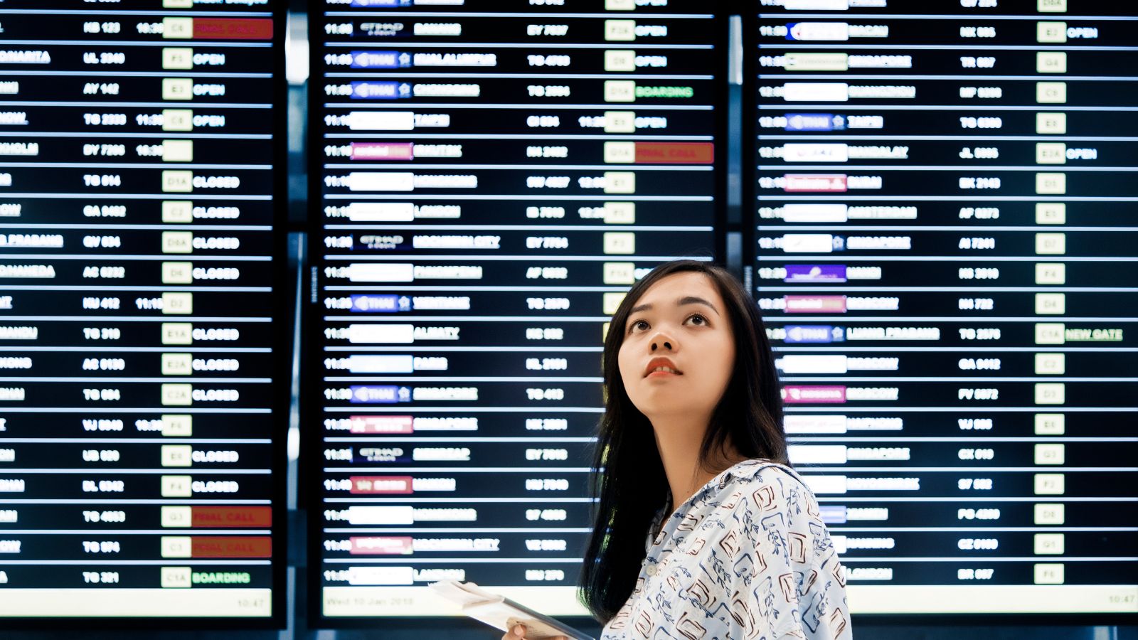A woman with documents looks up at a large airport departure board showing various flight times and statuses.