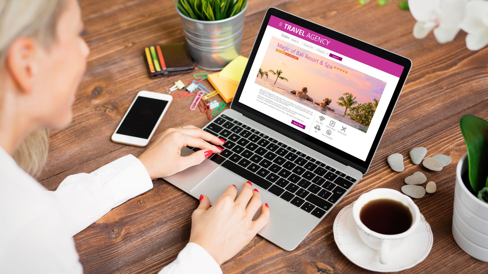 Woman browsing a travel agency website on her laptop at a wooden table with a phone, notepad, coffee cup, and small plant.