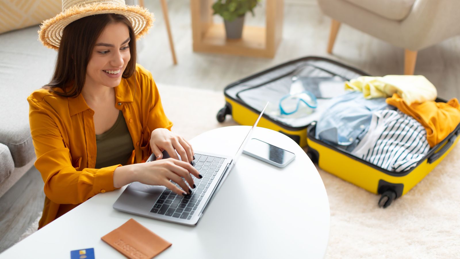 A woman in a hat uses a laptop at a table with a passport and phone, next to an open suitcase filled with clothes.