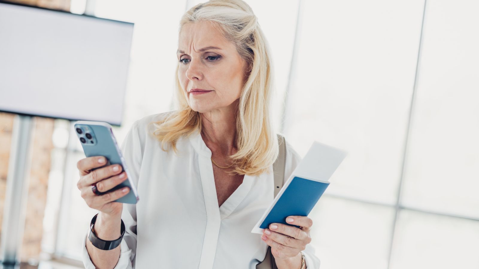 A woman indoors holds a smartphone and passport with documents, focusing intently on her phone.