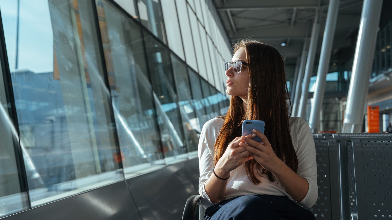 A woman with long brown hair and glasses sits in an airport terminal, holding her phone and gazing out at bright sunlight.