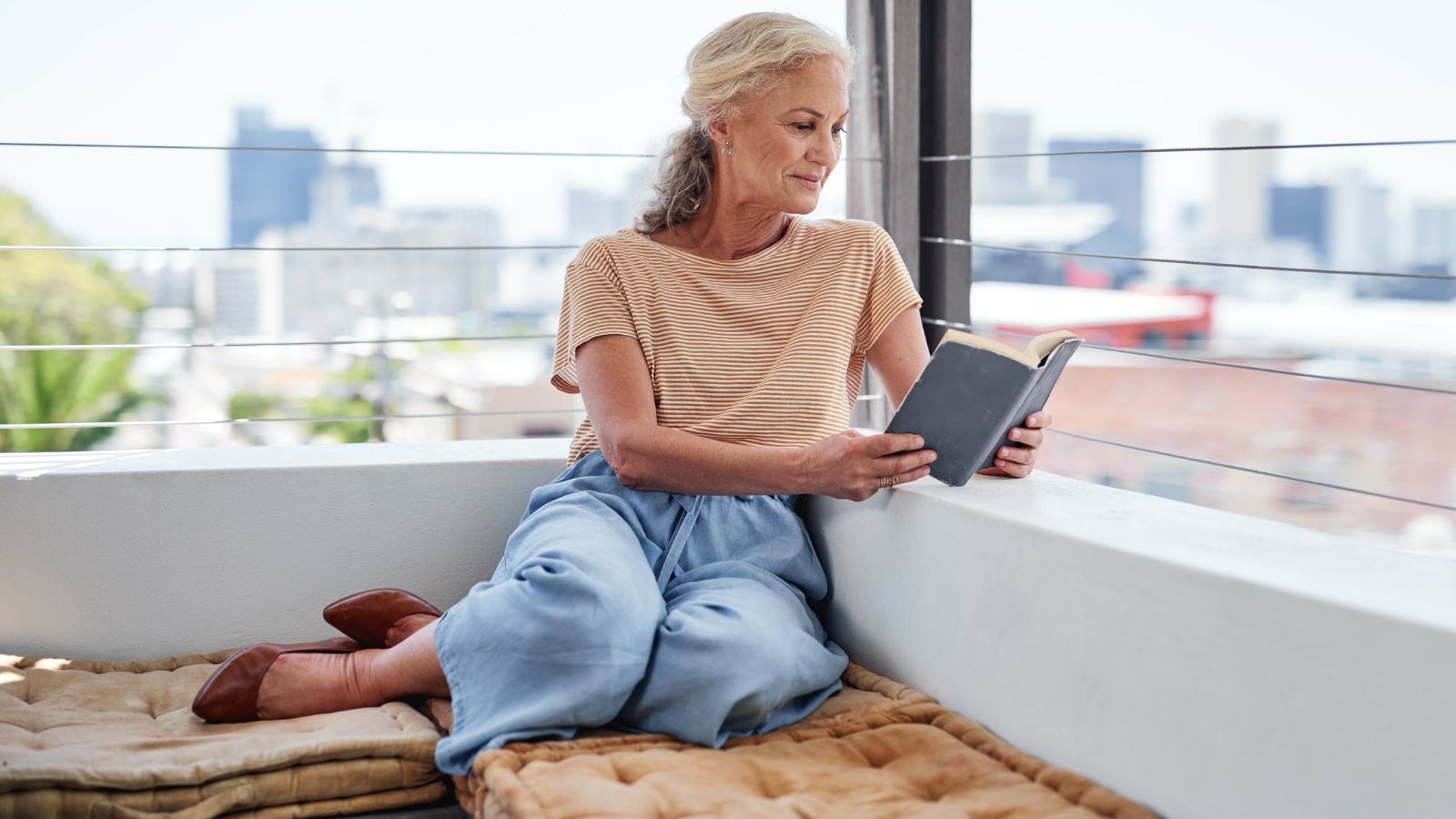 Older woman reads on a cushioned balcony bench with a city skyline in the background.