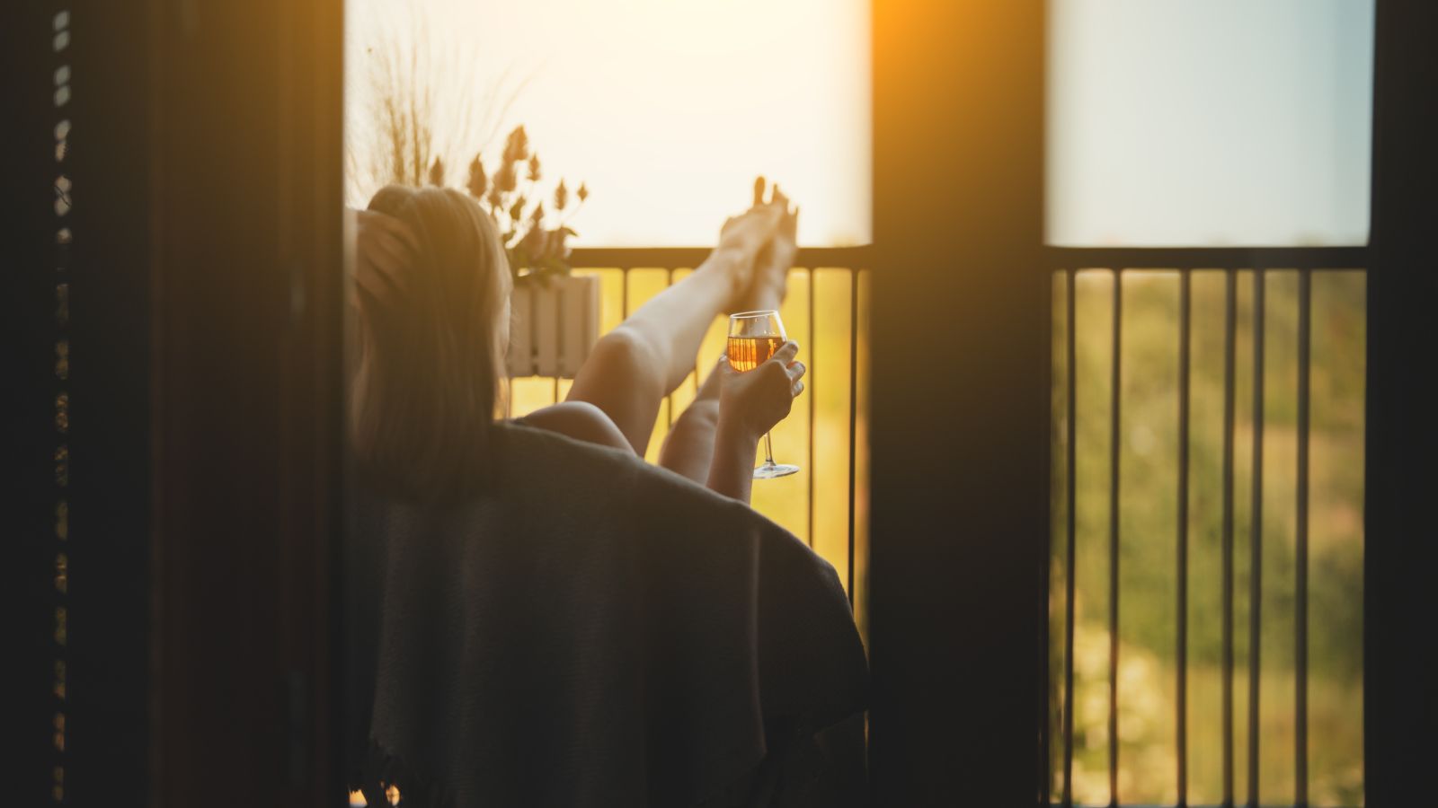 Person reclines on a balcony chair with feet up, holding a glass of wine and facing the sunset.