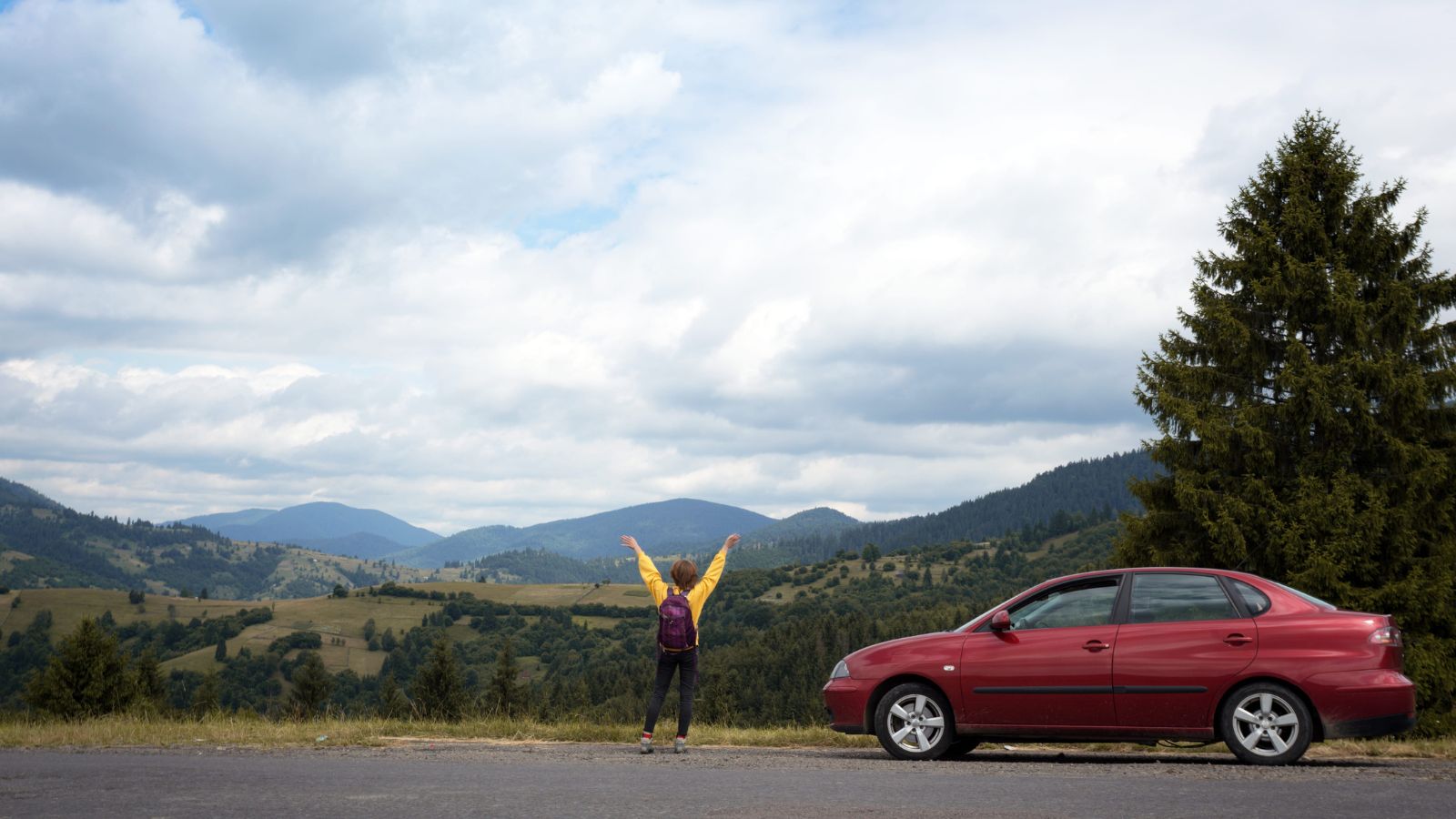 A photo of a driver stepping out of a parked car to stretch and walk at a scenic roadside area, mountains or countryside in background.