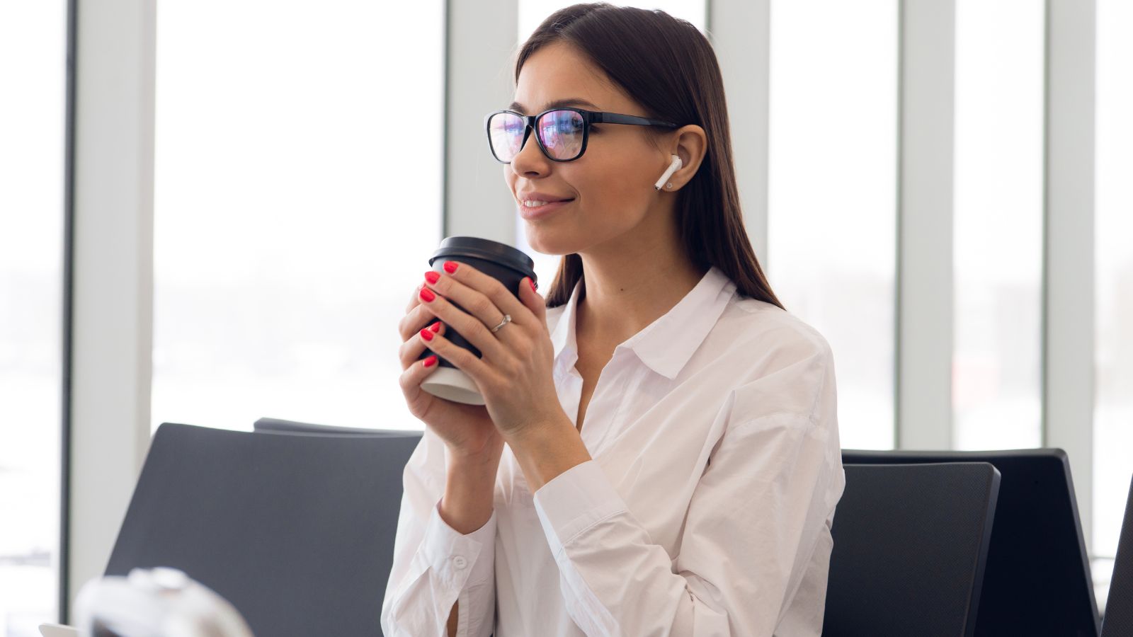 A woman with glasses and earbuds holds a coffee cup at a table in a bright room with large windows.
