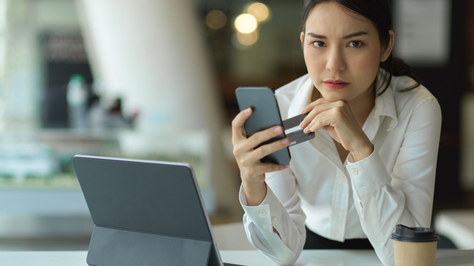 A serious-looking woman in a white shirt holds a phone and credit card, looking at a tablet on a desk with a coffee cup.